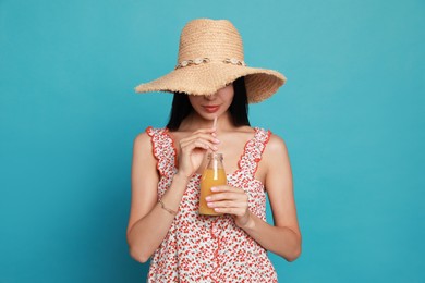 Beautiful young woman with straw hat and bottle of refreshing drink on light blue background