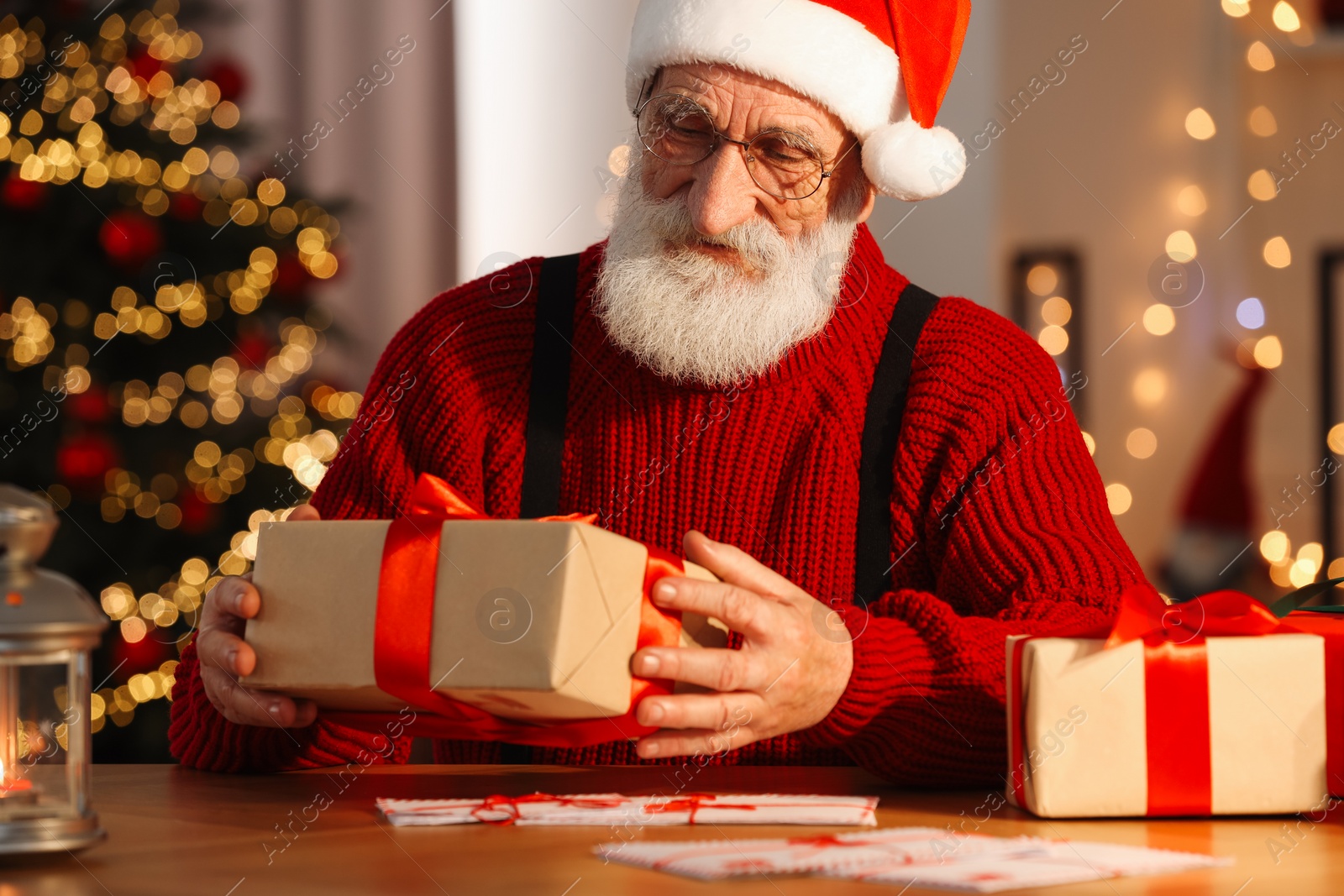 Photo of Santa Claus holding gift box at his workplace in room decorated for Christmas