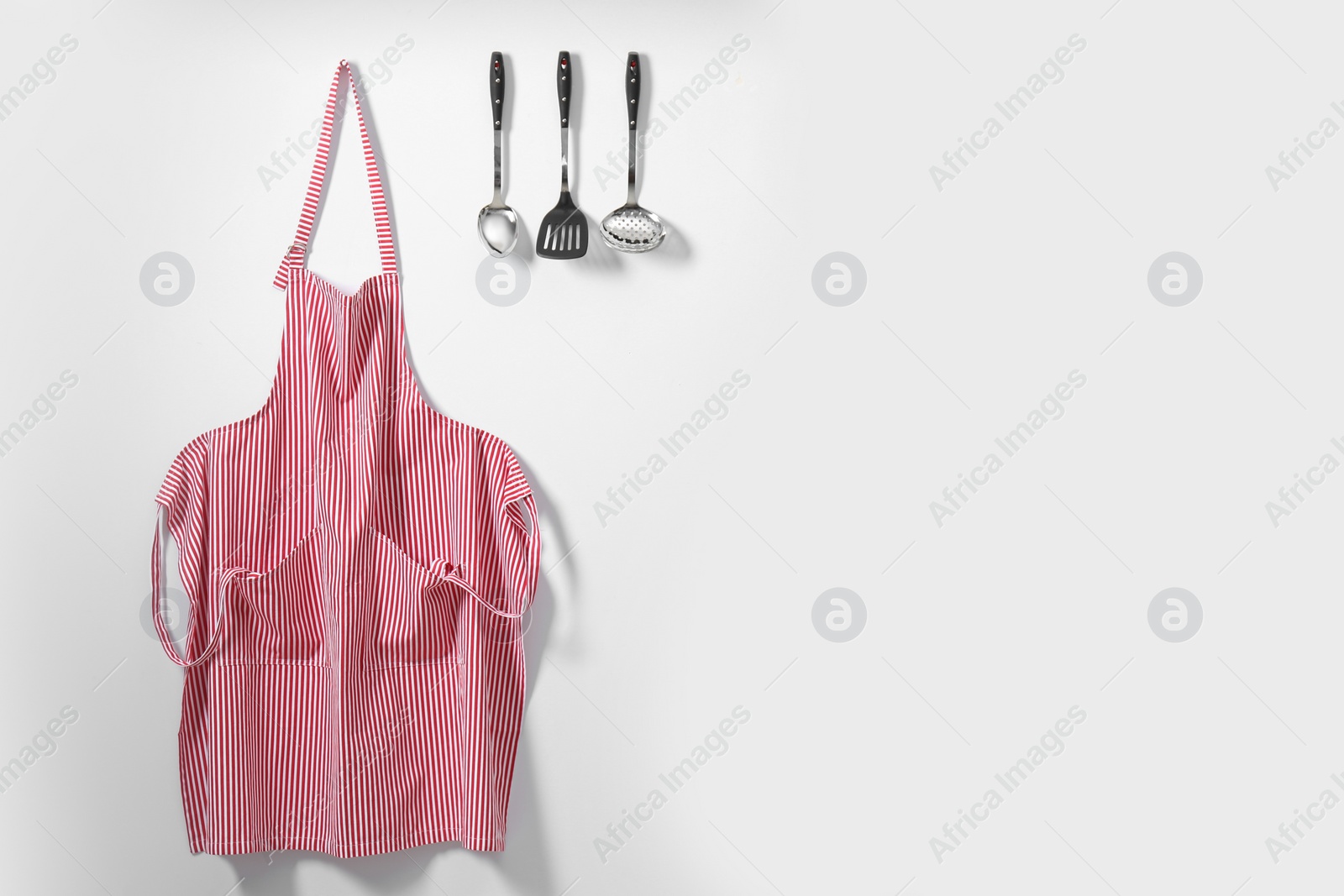 Photo of Red striped apron and kitchen tools on light wall. Space for text
