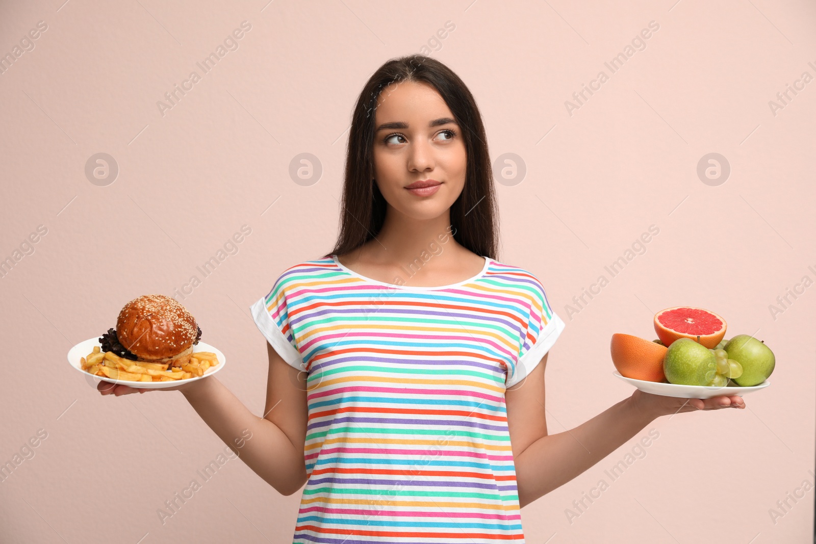 Photo of Woman choosing between fruits and burger with French fries on beige background