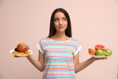 Woman choosing between fruits and burger with French fries on beige background