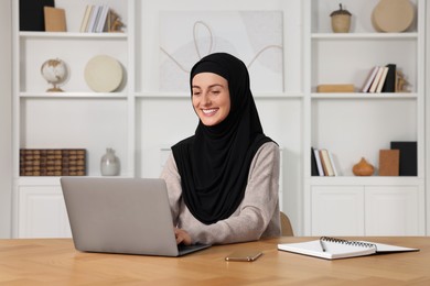 Photo of Muslim woman in hijab using laptop at wooden table in room