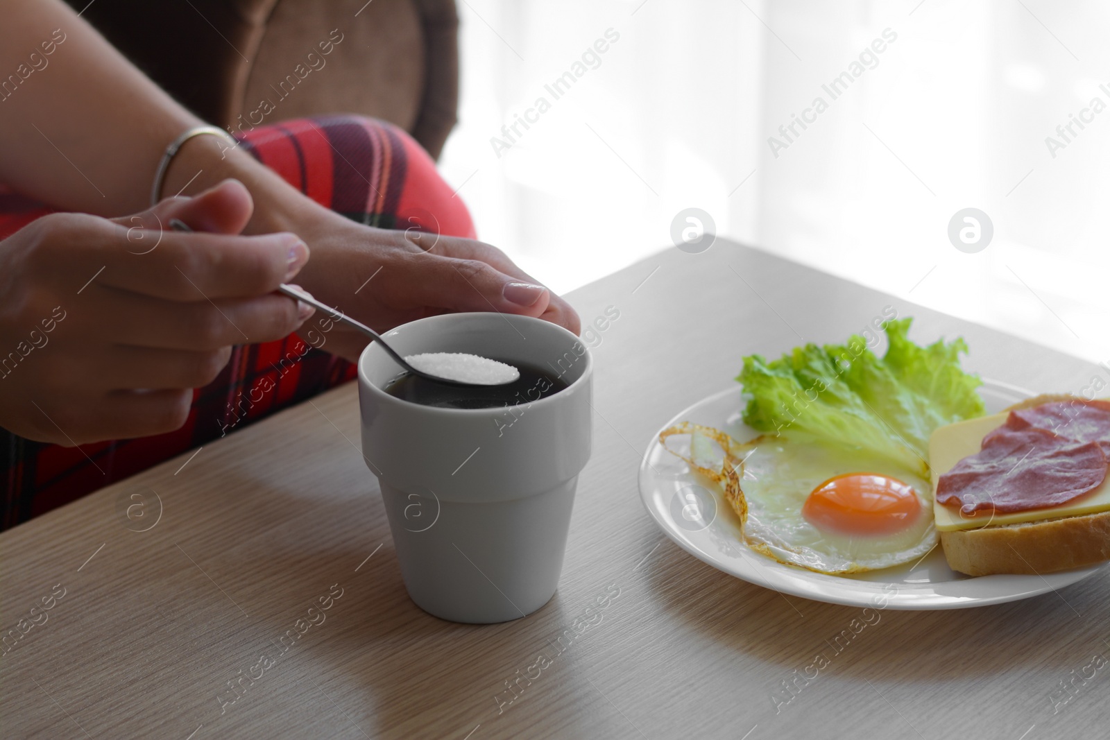 Photo of Woman adding sugar to morning coffee while having breakfast at wooden table indoors, closeup