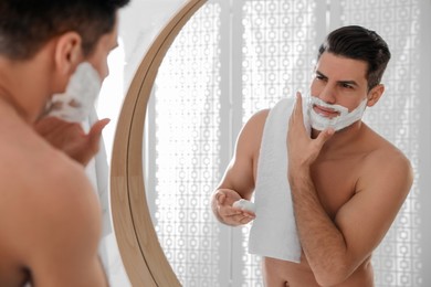 Handsome man applying shaving foam near mirror in bathroom