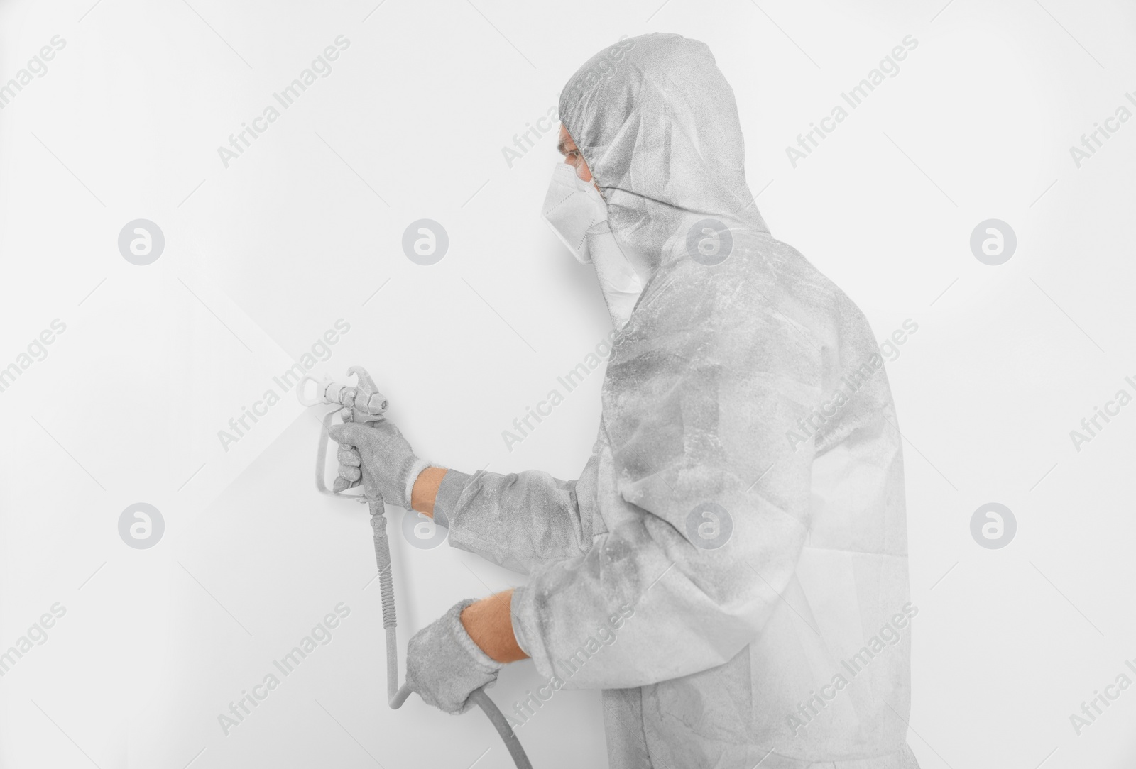 Photo of Decorator in protective overalls painting wall with spray gun indoors