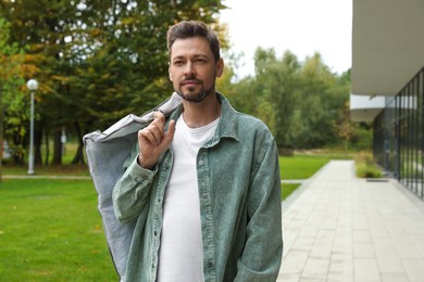 Photo of Attractive serious man holding garment cover with clothes outdoors. Dry cleaning service