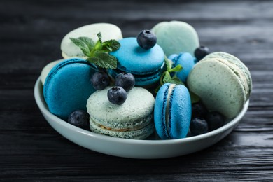 Photo of Delicious macarons, mint and blueberries in bowl on black wooden table, closeup