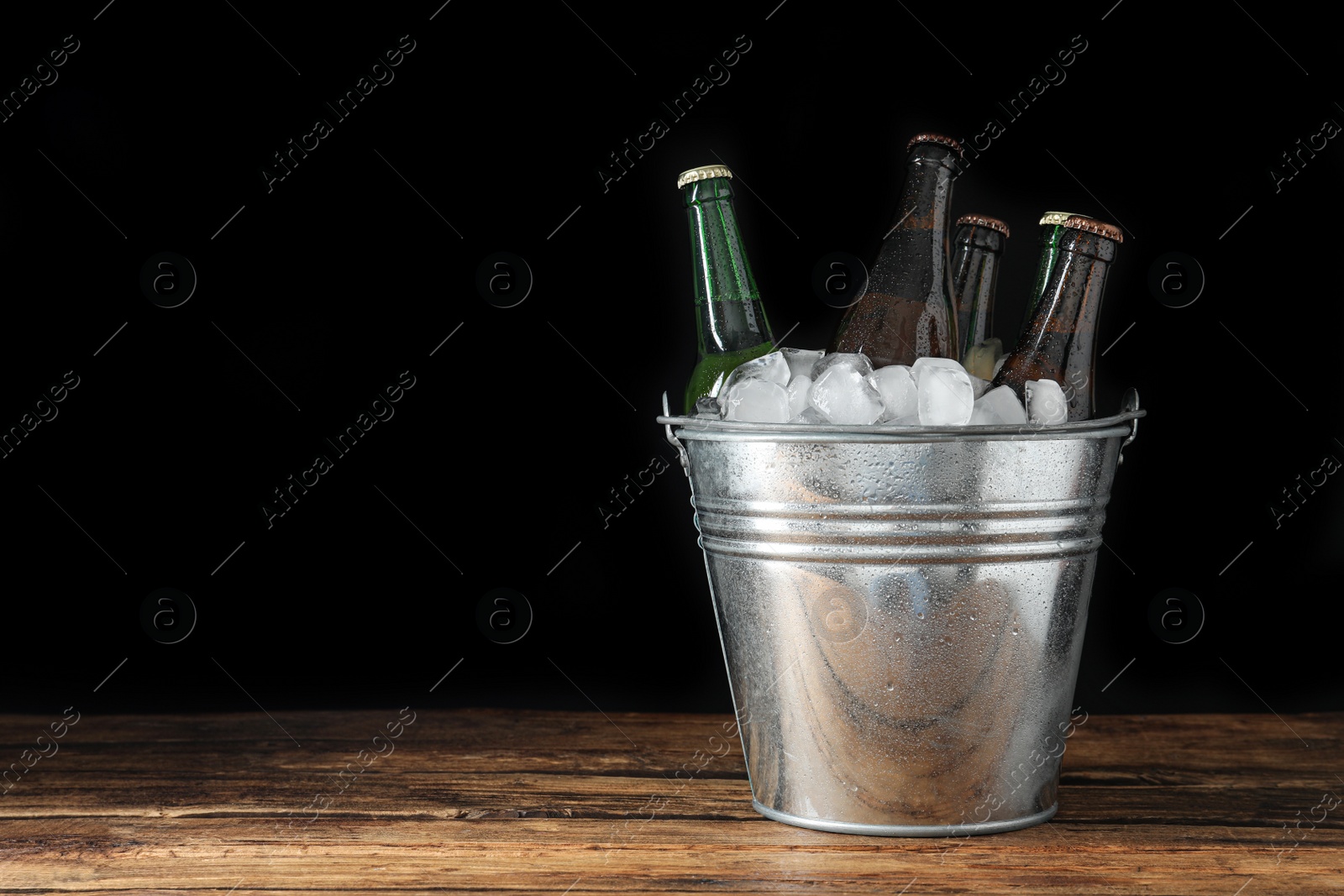 Photo of Metal bucket with bottles of beer and ice cubes on wooden table. Space for text