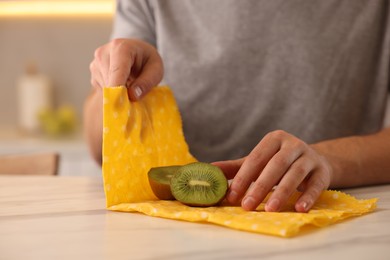 Photo of Man packing fresh kiwi into beeswax food wrap at light marble table in kitchen, closeup