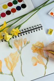 Photo of Woman painting freesias in sketchbook at white table, top view