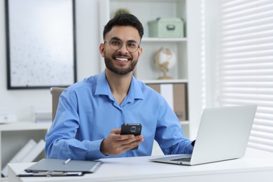 Photo of Happy young man using smartphone while working with laptop at white table in office