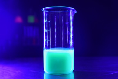 Laboratory beaker with luminous liquid on table against dark blue background, space for text