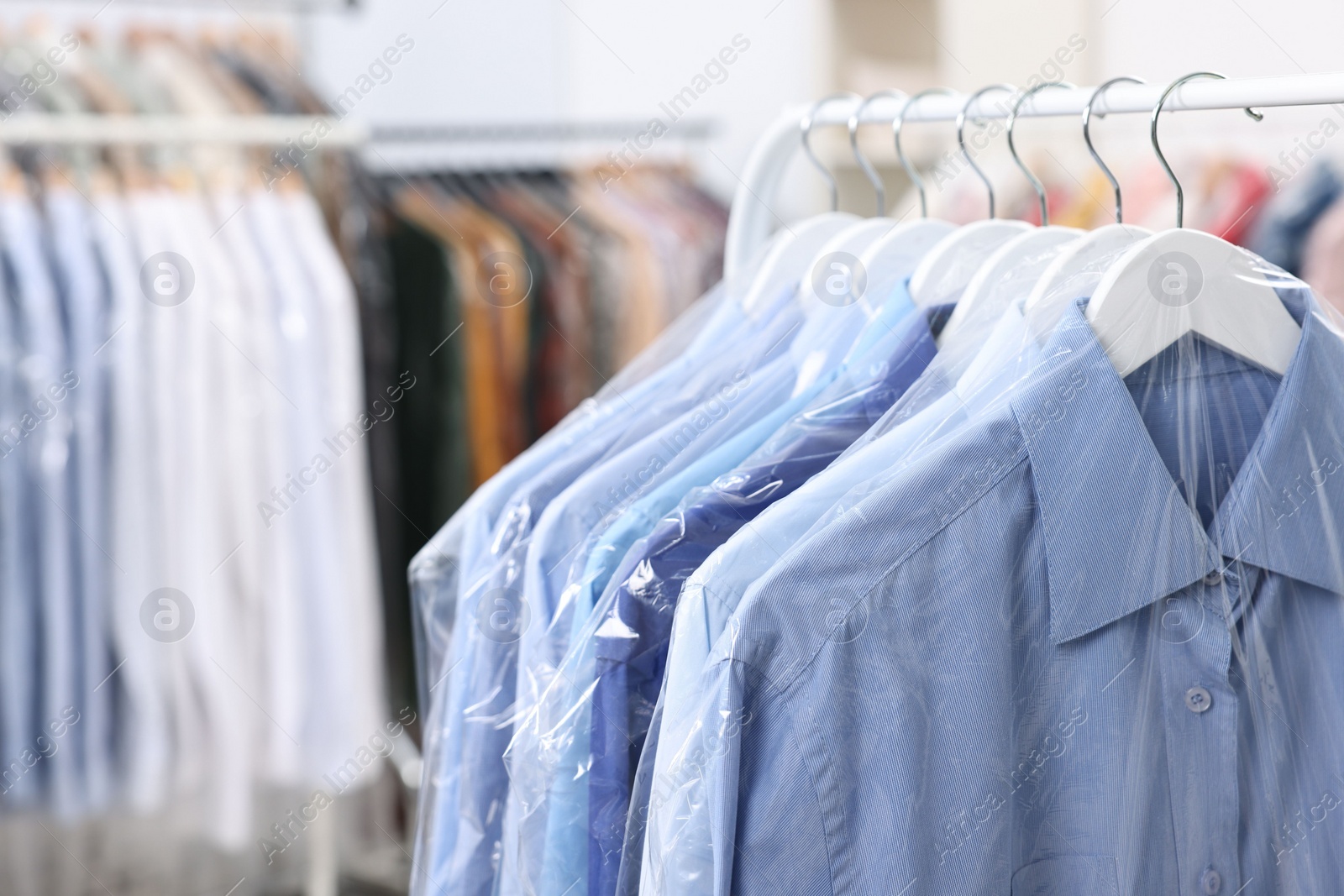 Photo of Dry-cleaning service. Many different clothes in plastic bags hanging on rack indoors, closeup