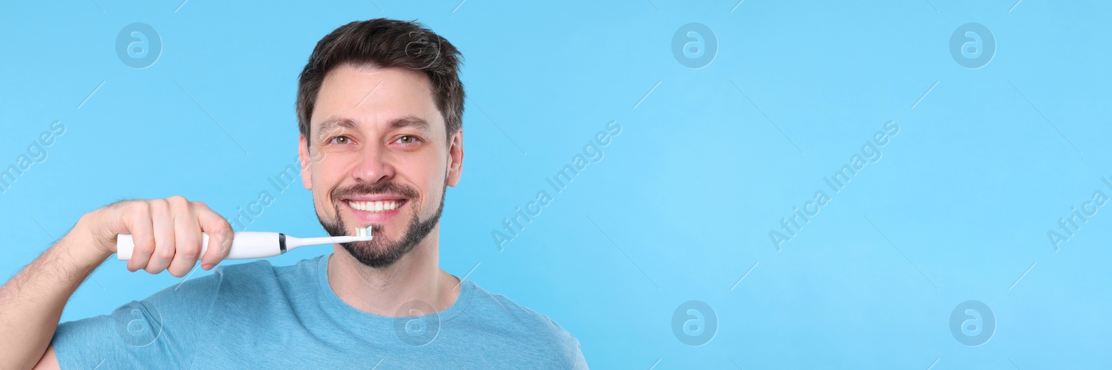 Image of Happy man using electric toothbrush on light blue background. Banner design with space for text