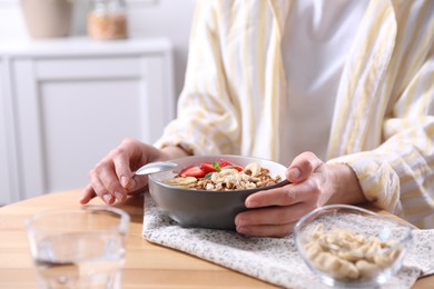 Photo of Woman eating tasty granola with banana, cashew and strawberries at wooden table indoors, closeup