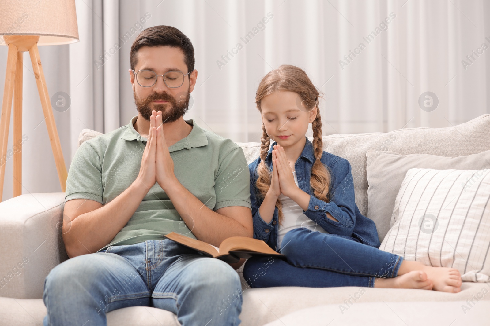 Photo of Girl and her godparent praying over Bible together on sofa at home
