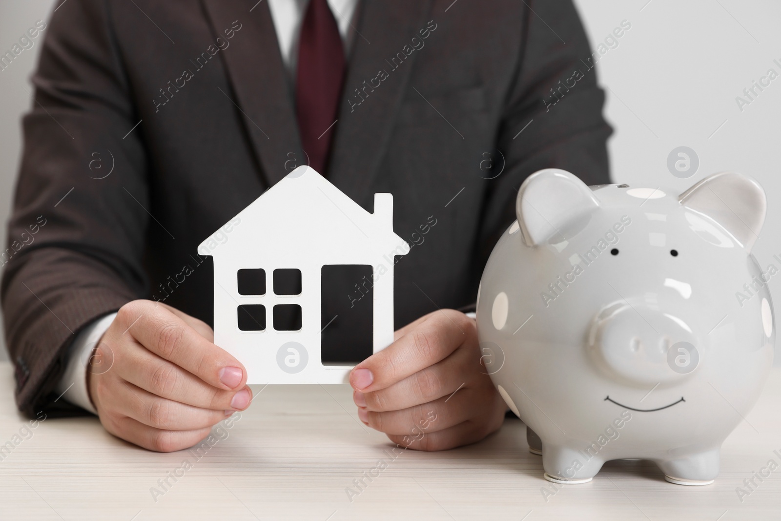 Photo of Man holding house model and piggy bank at wooden table, closeup. Saving money concept