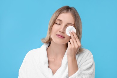 Young woman cleaning face with cotton pad on light blue background