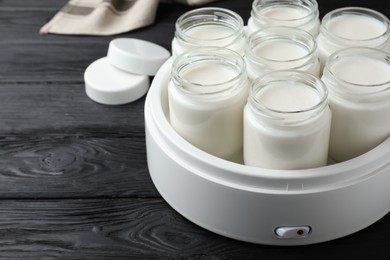 Photo of Modern yogurt maker with full jars on black wooden table. Space for text