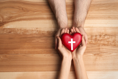 Couple holding heart with cross symbol on wooden background, top view with space for text. Christian religion