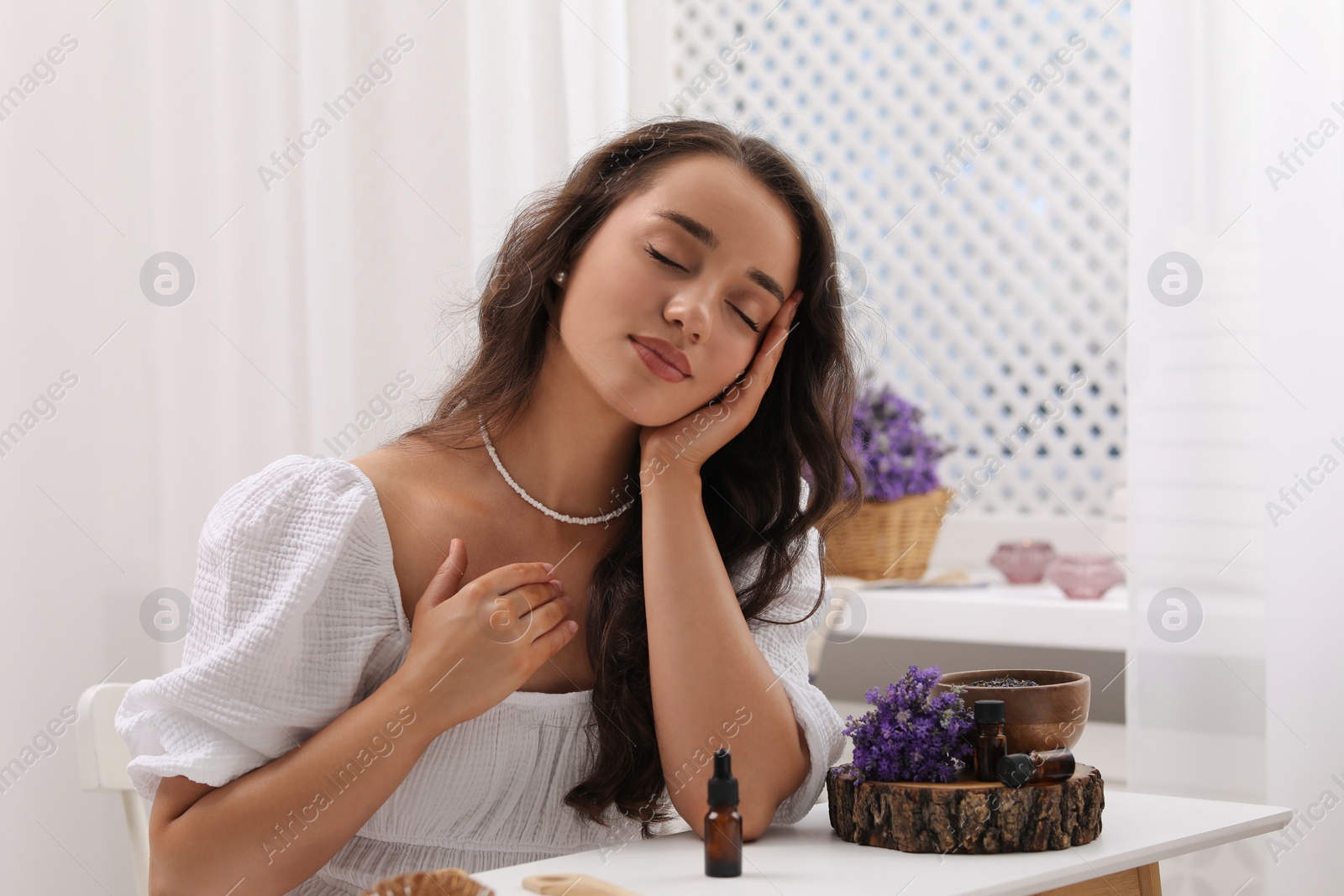 Photo of Beautiful young woman at table with bottles of essential oil and flowers indoors