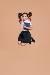 Cute schoolgirl holding book and jumping on beige background