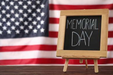Blackboard with phrase Memorial Day on wooden table against American flag, space for text