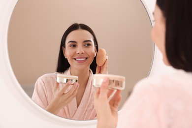 Photo of Beautiful young woman applying powder with puff applicator near mirror indoors