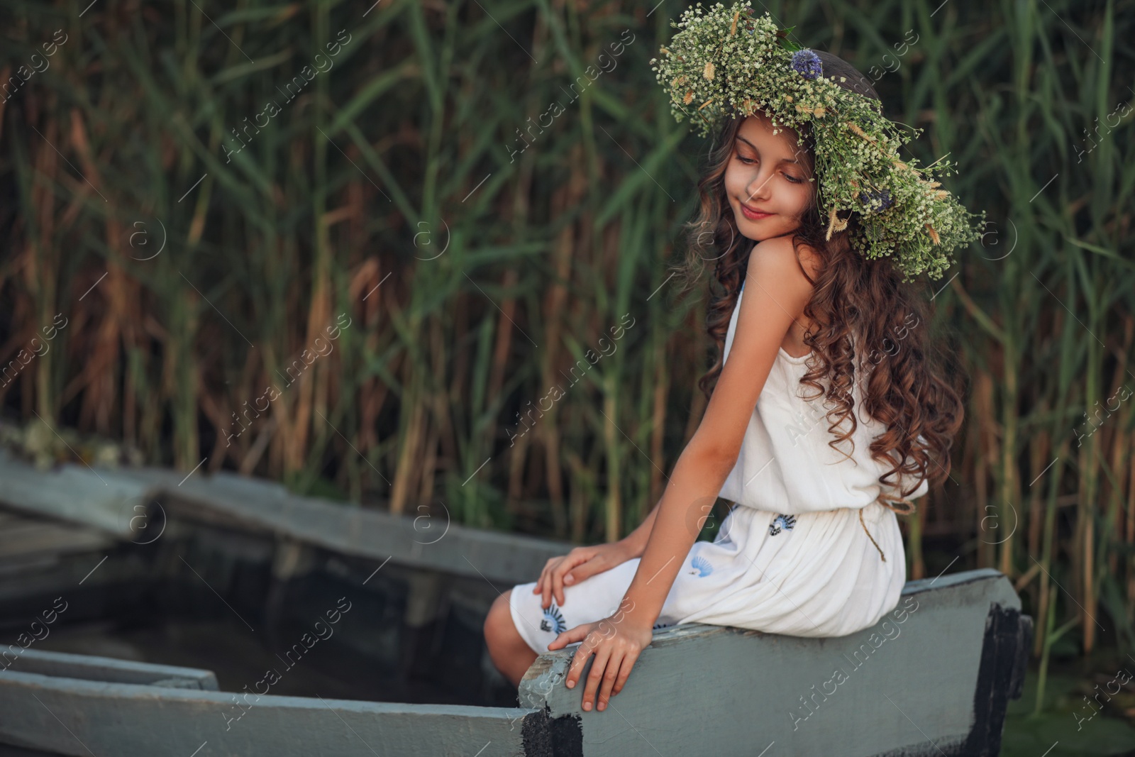 Photo of Cute little girl wearing wreath made of beautiful flowers in wooden boat