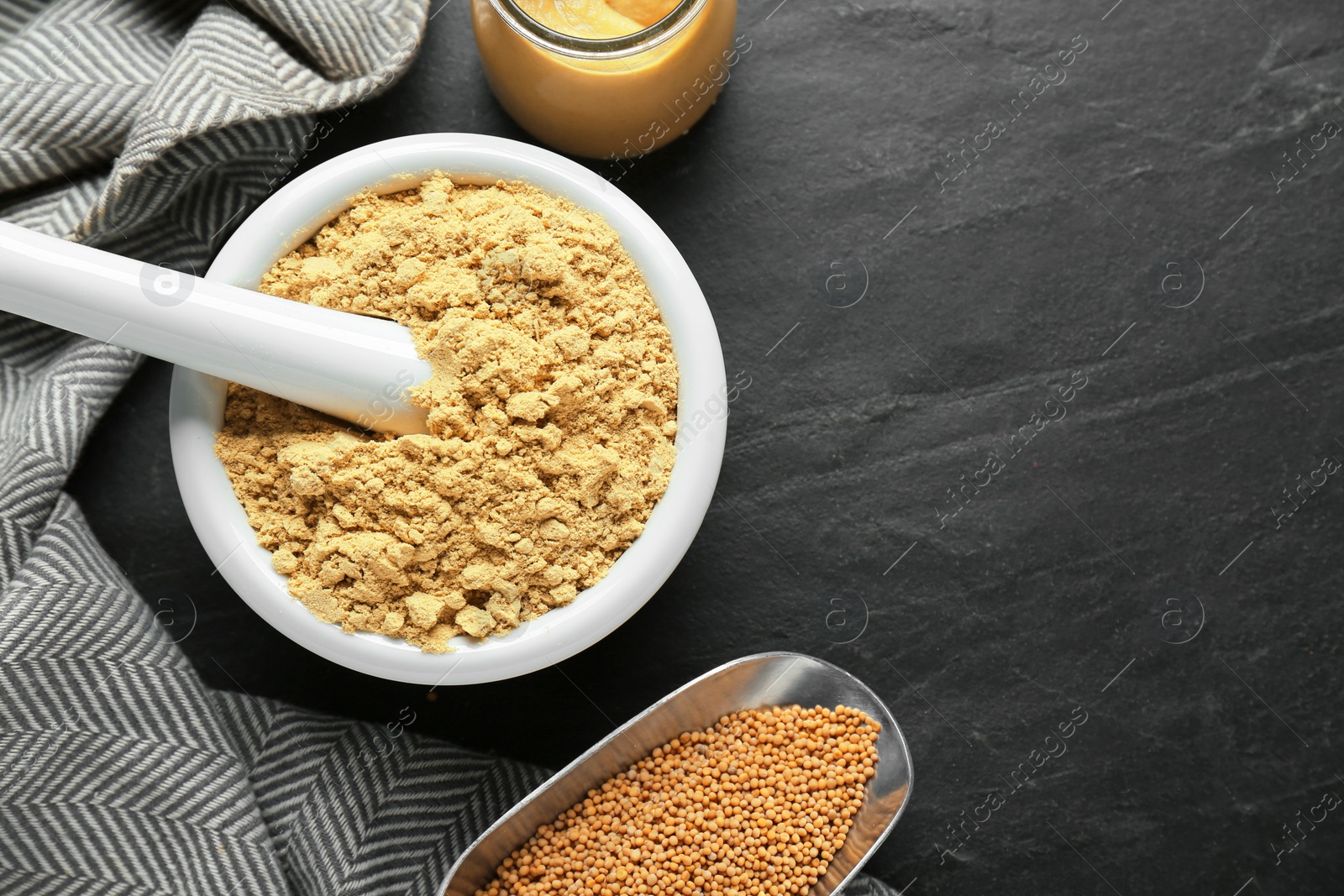 Photo of Mortar of mustard powder and scoop with seeds on black table, flat lay. Space for text