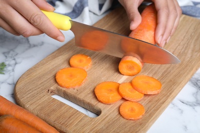 Woman cutting ripe carrot on wooden board at white marble table, closeup