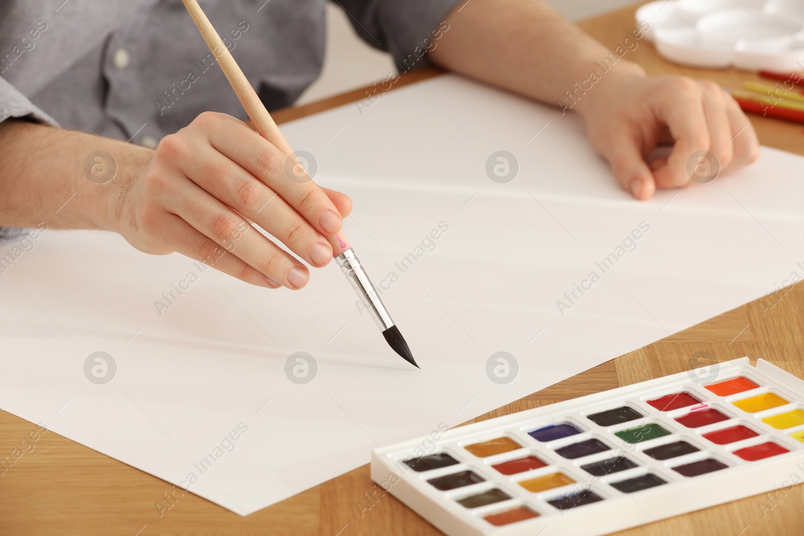 Photo of Man painting with watercolor on blank paper at wooden table, closeup