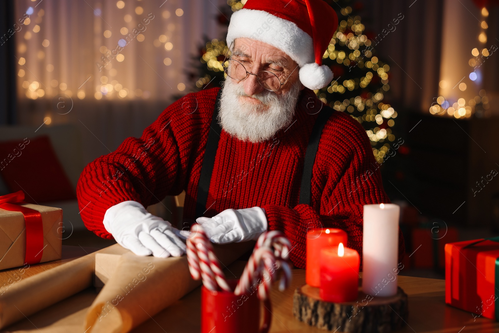 Photo of Santa Claus wrapping gift at his workplace in room decorated for Christmas