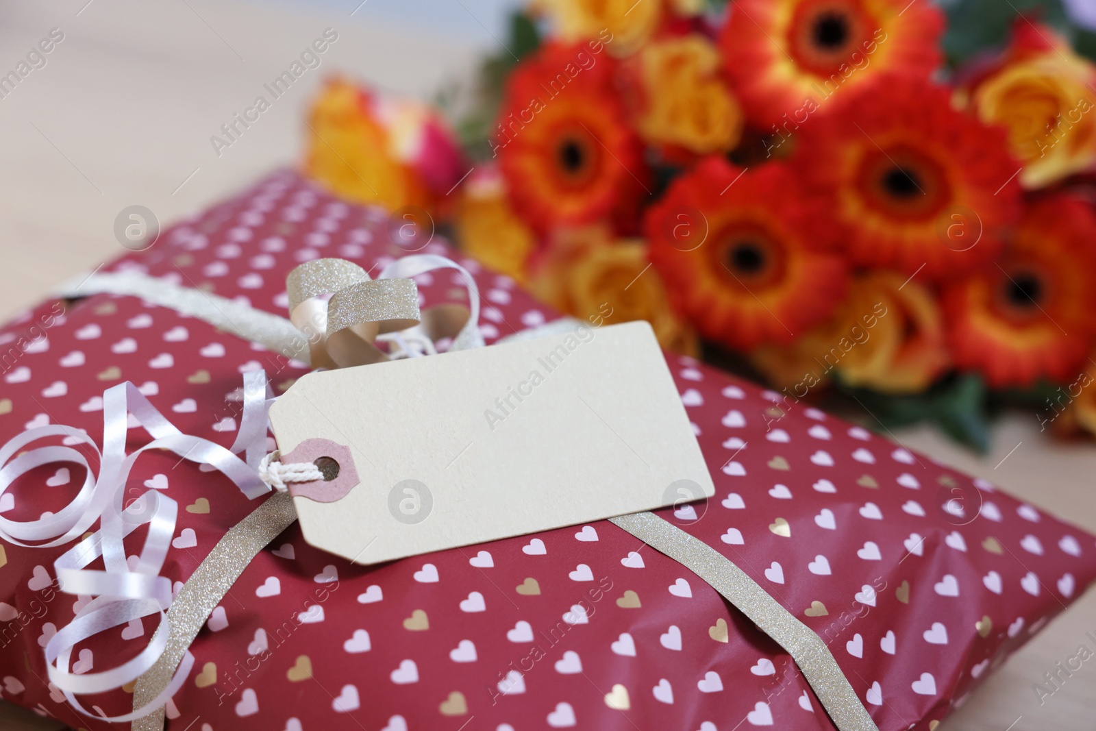 Photo of Parcel wrapped in heart patterned paper with cardboard tag and flowers on table, closeup. Space for text