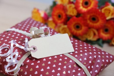 Photo of Parcel wrapped in heart patterned paper with cardboard tag and flowers on table, closeup. Space for text