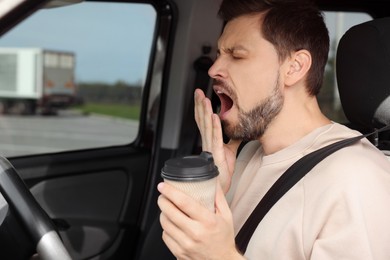 Sleepy man with cup of coffee yawning in modern car