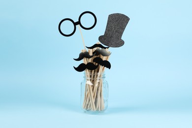 Photo of Fake paper mustaches, glasses, hat and straws in jar on light blue background