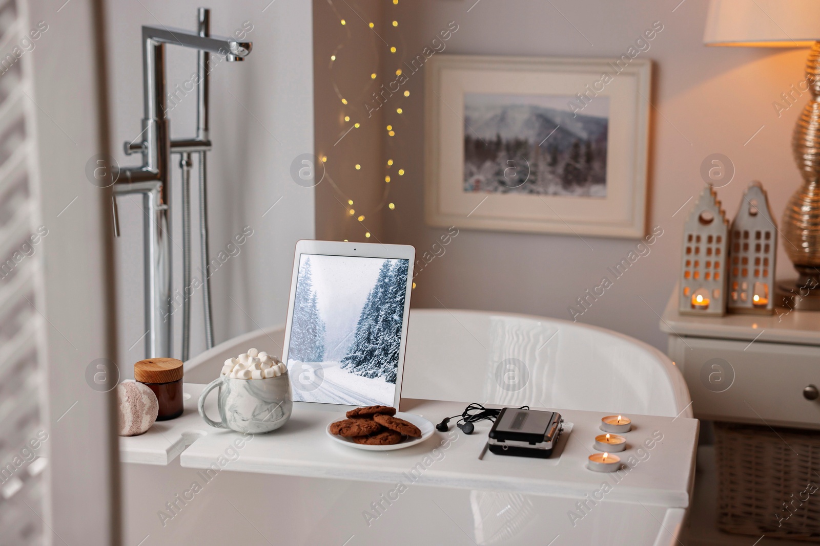 Photo of White wooden tray with tablet, spa products and burning candles on bathtub in bathroom