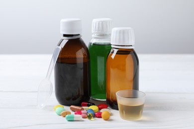 Photo of Bottles of syrup, measuring cup, dosing spoon and pills on white table. Cold medicine