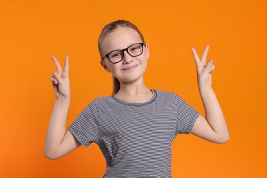 Photo of Portrait of cute girl in glasses showing victory gesture on orange background