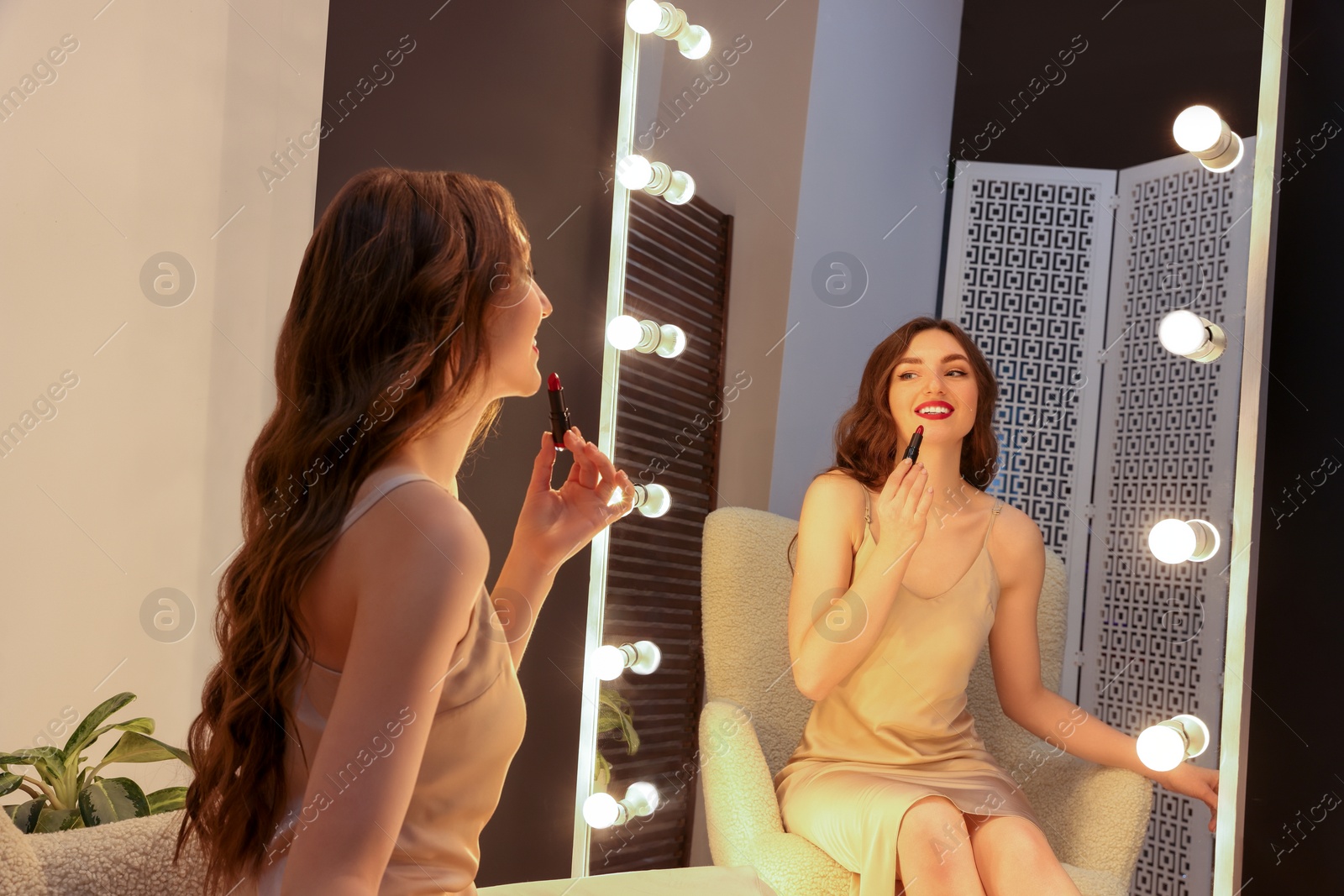 Photo of Beautiful young woman in elegant dress applying lipstick near mirror indoors