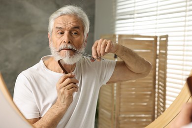 Photo of Senior man trimming mustache with scissors near mirror in bathroom