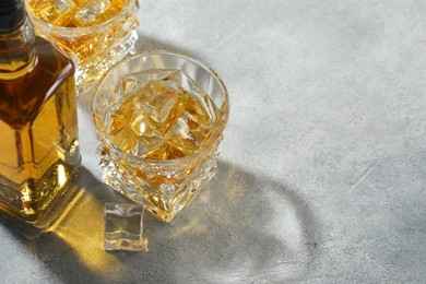 Whiskey with ice cubes in glasses and bottle on grey textured table, space for text