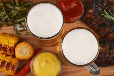 Photo of Mugs with beer, delicious grilled ribs and ingredients on wooden table, closeup