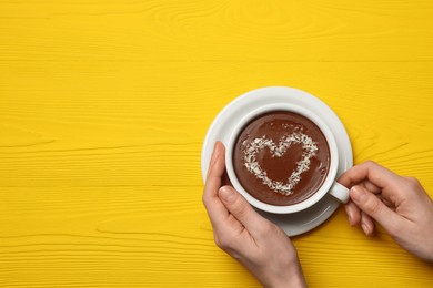 Photo of Woman holding cup of hot chocolate with heart shaped decoration at yellow wooden table, top view. Space for text