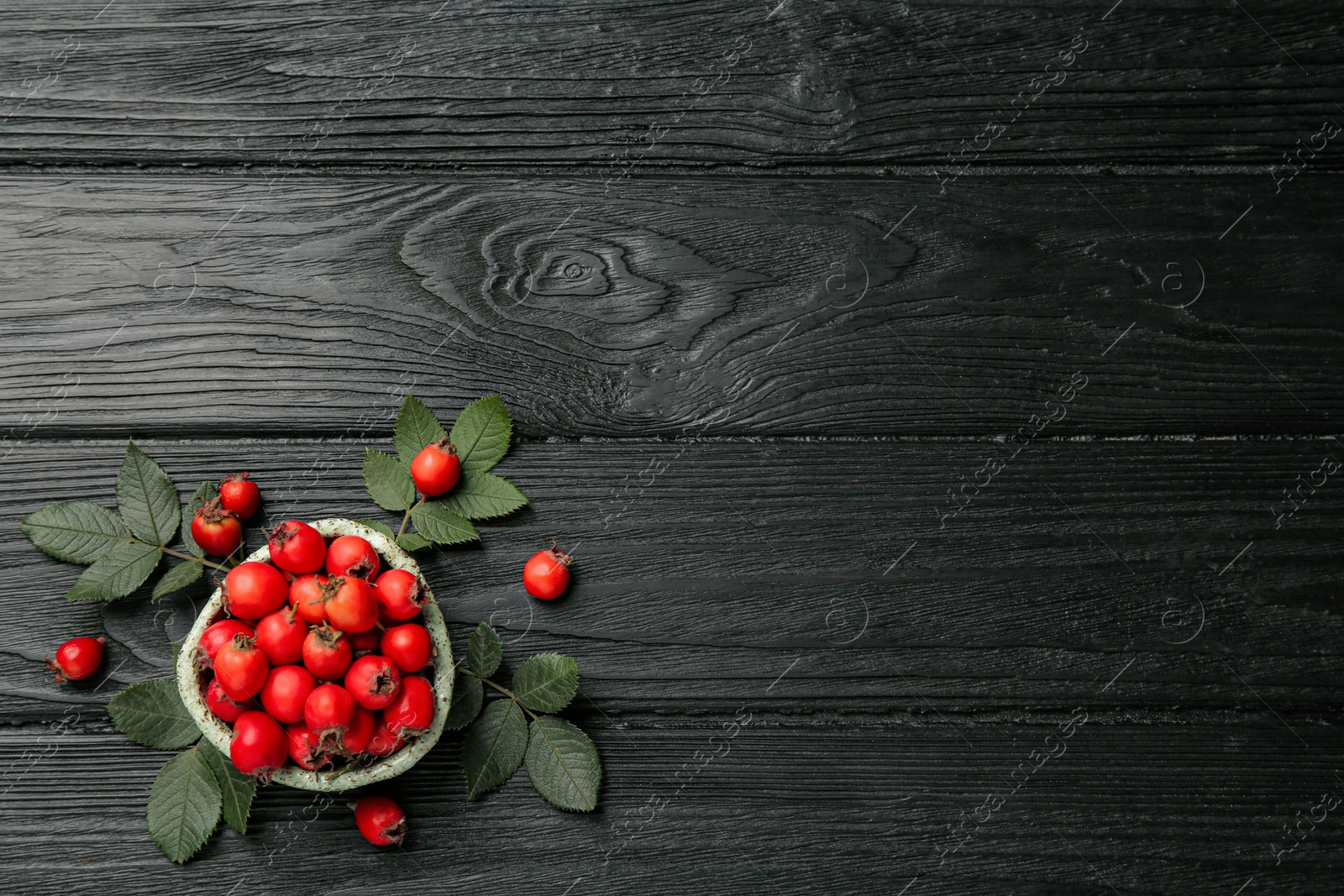 Photo of Ripe rose hip berries with green leaves on black wooden table, flat lay. Space for text