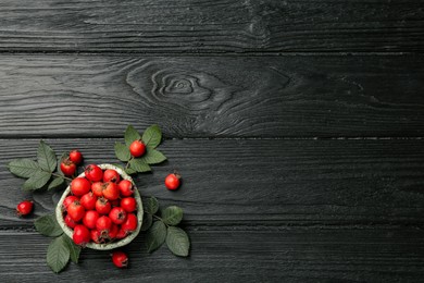 Ripe rose hip berries with green leaves on black wooden table, flat lay. Space for text