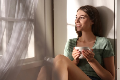 Beautiful young woman with cup of drink near window at home