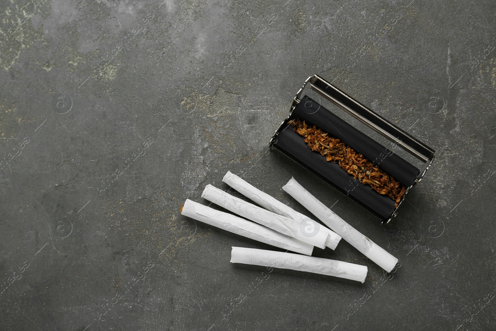 Photo of Roller with tobacco and hand rolled cigarettes on dark grey table, flat lay. Space for text