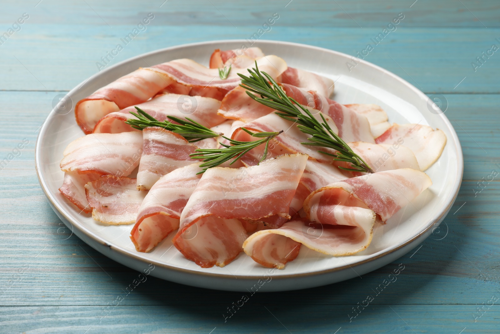 Photo of Slices of raw bacon and rosemary on light blue wooden table, closeup